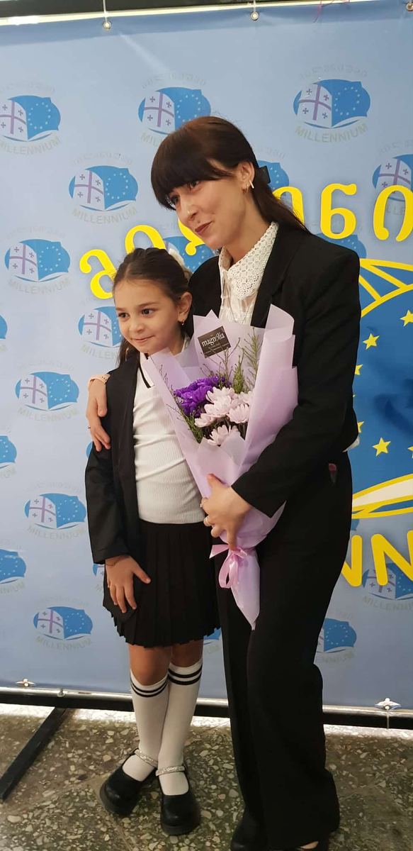 მასწავლებელი და პატარა მოსწავლე ყვავილების თაიგულით სკოლის ბანერთან